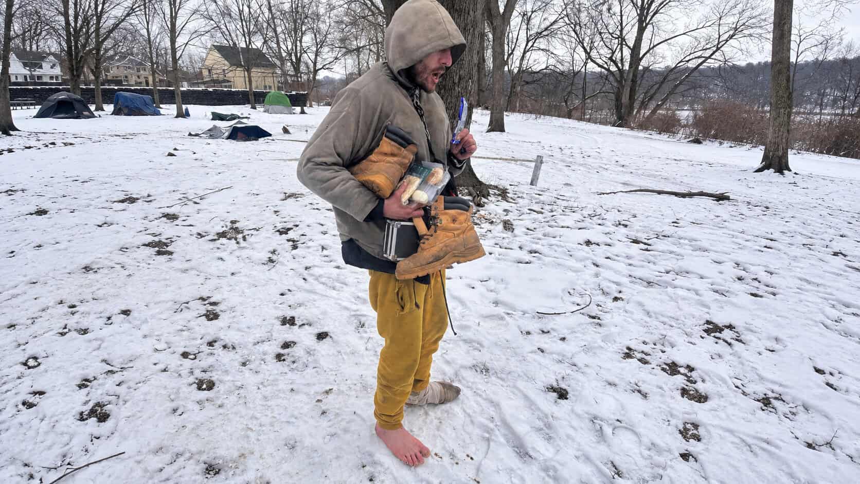Aaron standing in the snow with one bare foot and one wrapped foot, holding his work boots and food.